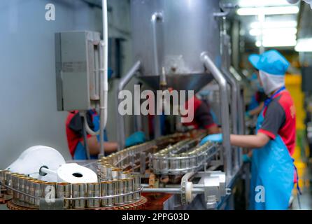 Canned fish factory. Food industry.  Sardines in red tomato sauce in tinned cans on conveyor belt at food factory. Blur workers working in food processing production line. Food manufacturing industry. Stock Photo