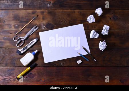 A school or office still life with a white blank sheet of paper and many office supplies. The school supplies lie on a brown wooden background. Place for text Stock Photo