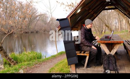 Man work on laptop charging from portable solar panel utdoor in alclove near the river. Clean energy for using in camping. Work with gadgets outdoor with portable solar panels. Stock Photo