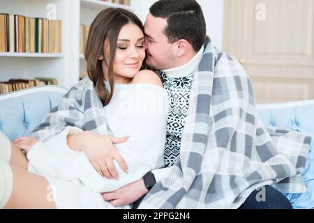 couple is hugging while sitting on a sofa and covered with a blanket Stock Photo
