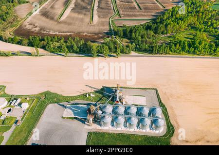 Aerial View Modern Granary, Grain-drying Complex, Commercial Grain Or Seed Silos In Sunny Spring Rural Landscape. Corn Dryer Silos, Inland Grain Terminal, Grain Elevators Standing In A Field Stock Photo