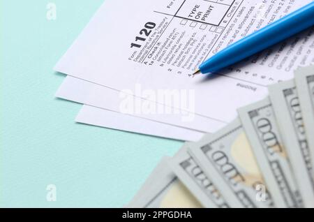 1120 tax form lies near hundred dollar bills and blue pen on a light blue background. US Corporation income tax return Stock Photo