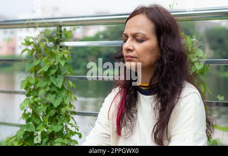 Active Mid Adult Woman in meditation sitting with eyes closed concentrate in a public park against garden leaves in the background. Front view. Head and Shoulder Shot. Close up Portrait. Stock Photo