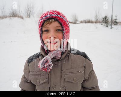 A cute boy 8 years old in a knitted red hat with braids and tassels. The child smiles cheerfully. Snowflakes on the face. Winter holidays, frosty weather and travel in the north Stock Photo