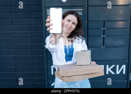 Happy woman holding mobile phone with white screen mockup for inscription and holding many different cardboard boxes with online order, satisfied with fast delivery, buying high quality products. Stock Photo