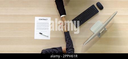 Asian Human Resource Manager in black suit shake hands to congratulate young candidate after the interview. Computer monitor, keyboard, mouse and contract document are on wooden table. Top View Stock Photo