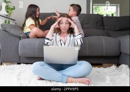 Tired busy woman working on laptop at home with her children. Single mother telecommuting stress. Stock Photo
