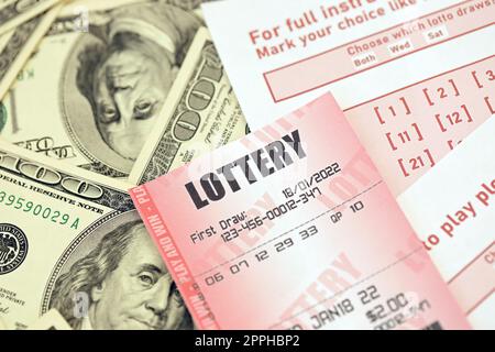 Red lottery ticket lies on pink gambling sheets with numbers for marking on big amount of hundred dollar bills. Lottery playing concept or gambling addiction. Close up Stock Photo