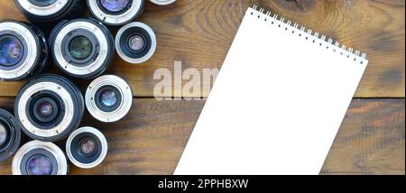 Several photographic lenses and white notebook lie on a brown wooden background. Space for text Stock Photo