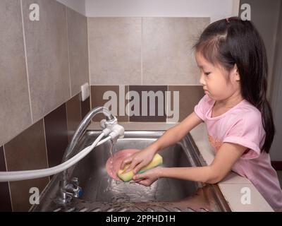 Adorable  kid girl washing dishes in domestic kitchen. Child having fun with helping his parents with housework. Stock Photo