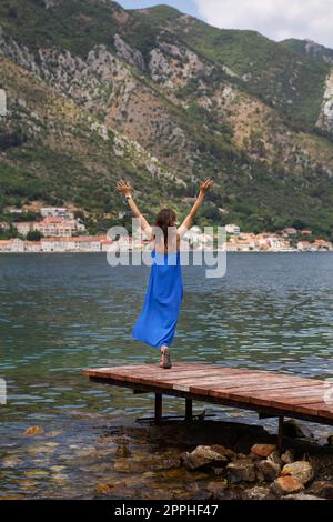 A girl in a blue dress stands with her arms raised on a pier against the background of the Bay of Kotor in Montenegro. A beautiful and cozy old town. Stock Photo