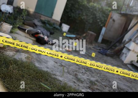 Victim of a violent crime in a backyard of residental house in evening. Dead man body under the yellow police line tape and evidence markers on crime scene Stock Photo