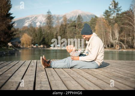 Happy family. Father playing with her baby boy infant oudoors on sunny autumn day. Portrait of dad and little son on wooden platform by lake. Positive human emotions, feelings, joy. Stock Photo