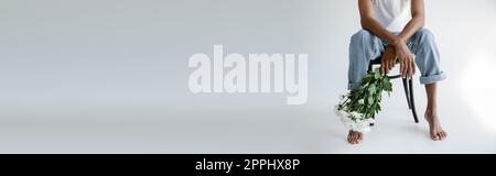 partial view of barefoot african american man in jeans sitting on chair with bouquet on grey background, banner,stock image Stock Photo