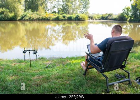 A man sits in a chair near the autumn lake and works on a laptop freelance camping. Robot out of the office, phoned with colleagues. Stock Photo