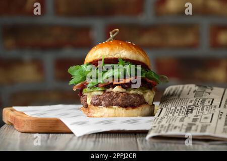 Burger on the wooden table with cheese, bacon, tomatos near newspaper. Stock Photo
