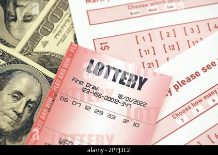 Red lottery ticket lies on pink gambling sheets with numbers for marking on big amount of hundred dollar bills. Lottery playing concept or gambling addiction. Close up Stock Photo