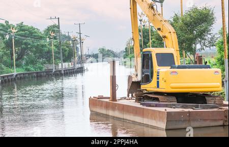 A yellow backhoe parked on a raft after dredging a canal. Backhoe on a pontoon and clean water in the river. Clean environment. Dredge aquatic plants from the canals with a backhoe to keep them clean. Stock Photo