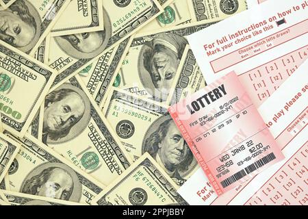 Red lottery ticket lies on pink gambling sheets with numbers for marking on big amount of hundred dollar bills. Lottery playing concept or gambling addiction. Close up Stock Photo