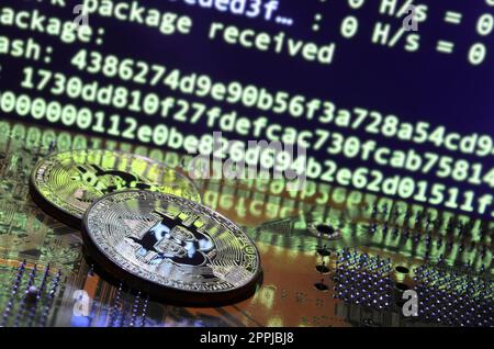 Two bitcoins lies on a videocard surface with background of screen display of cryptocurrency mining by using the GPUs Stock Photo