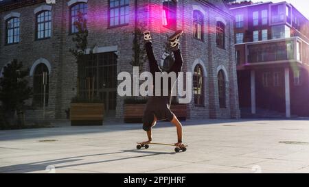 Agile Professional Skater Stands on the Hands on His Skateboard. Strong Guy Doing Handstand Tricks on a Longboard. In the Background Fashionable Hipst Stock Photo