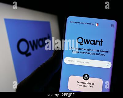 Person holding cellphone with website of French search engine company Qwant SAS on screen in front of logo. Focus on center of phone display. Stock Photo