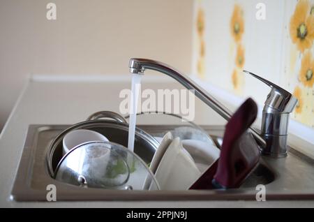Dirty dishes and unwashed kitchen appliances lie in foam water under a tap from a kitchen faucet Stock Photo