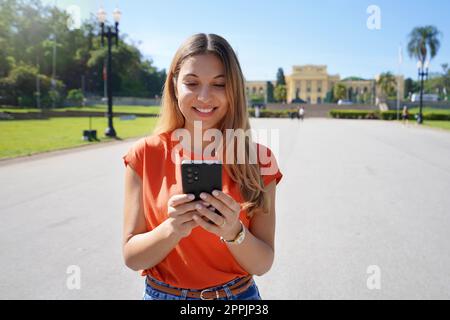 Brazilian young woman laughing and texting on her mobile phone walking outdoors on sunny day Stock Photo