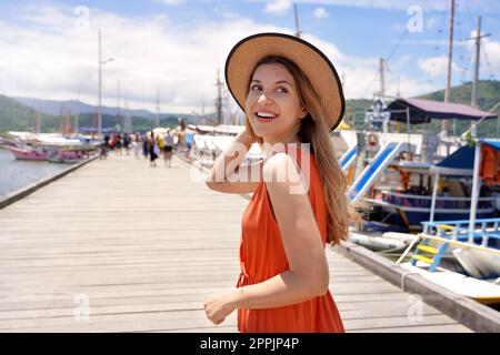 Portrait of smiling tourist woman walking on wooden pier where travel by ferry or rented boat or yacht to explore warm tropical destinations Stock Photo