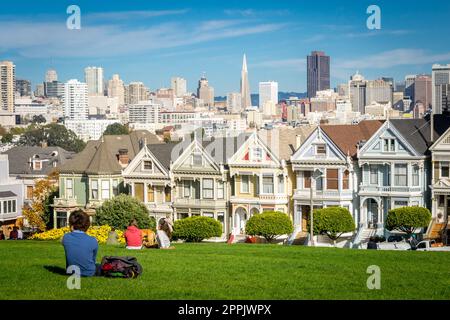 View on the Painted Ladies Victorian houses of San Francisco with cityscape and skyline in the background on a blue sky. People sitting front Stock Photo