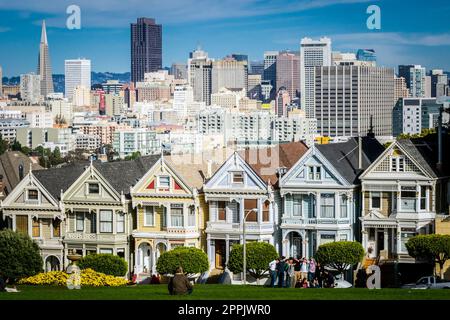 View on the Painted Ladies Victorian houses of San Francisco with cityscape and skyline in the background on a blue sky. People sitting front Stock Photo
