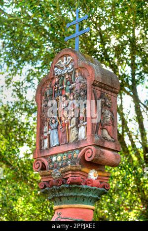 A wayside shrine colorfully decorated with Christian symbols and figures of saints in front of the green foliage of a tree Stock Photo