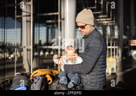 Fatherat comforting his crying infant baby boy child tired sitting on top of luggage cart in front of airport terminal station while traveling wih family. Stock Photo