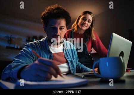 Young serious man calculating monthly expenses, writing in a notebook the bank numbers that he reads on the laptop, in the background, his wife watching him. Couple taking care of their finances. High quality photo Stock Photo