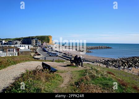 Elevated view across the town rooftops towards the beach, cliffs and countryside with a couple sitting on a bench in the foreground, West Bay, Dorset. Stock Photo