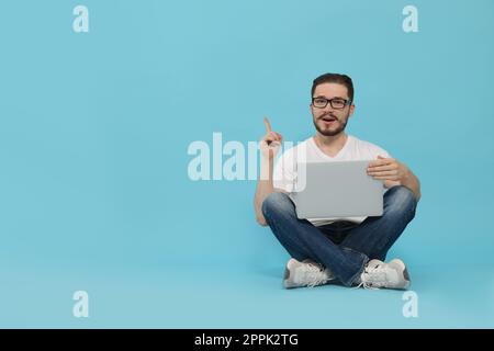 Emotional man sitting with laptop and pointing at something on light blue background. Space for text Stock Photo