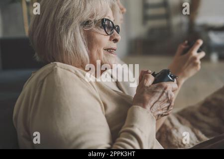 Emotional old woman having video games competition at home with millennial adult daughter sitting on sofa in living room holding joysticks. Stock Photo