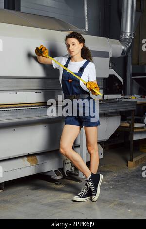 Apprentice woman leaned against a woodworking machine with a tape measure in hand Stock Photo