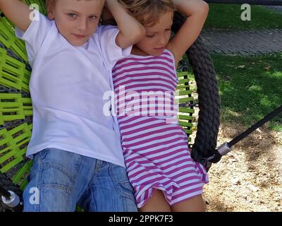 Children swing on a spider-web swing. Boy and girl, brother and sister in the summer on the playground. Swinging the swing. Boy 8 years old in a white T-shirt. Girl 6 years old in a striped pink dress Stock Photo