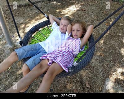 Children swing on a spider-web swing. Boy and girl, brother and sister in the summer on the playground. Swinging the swing. Boy 8 years old in a white shirt. Girl 6 years old in a striped pink dress Stock Photo