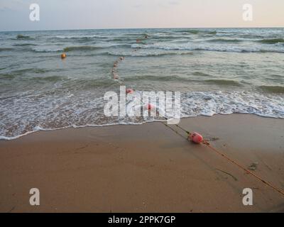 Buoys on a rope near sea water. The buoys are pink restraints to alert people to the depth of the water. Rescue of the drowning. Delimiting a place on a sandy beach between hotels. Wave with bubbles Stock Photo