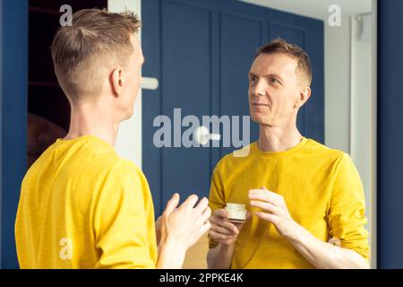 Happy mature man hold jar of moisturizing cream and apply on clear face skin. Male portrait in room mirror. Stock Photo