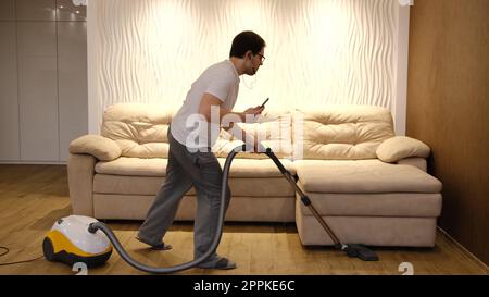 Young caucasian man in white and gray clothes having fun and dancing while vacuuming at home with biege sofa on background. Man in earphones with phone in hand have dancing with vacuum cleaner Stock Photo
