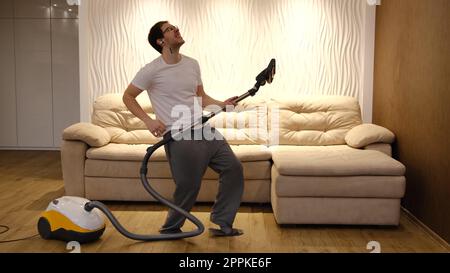 Young caucasian man in white and gray clothes having fun and dancing while vacuuming at home with biege sofa on background. Man in earphones have dancing with vacuum cleaner Stock Photo