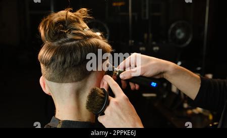 Shot of a handsome barber giving a haircut to his client using trimmer. Hairdresser service in a modern barbershop in a dark key lightning with warm light back view Stock Photo