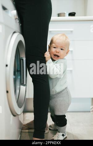 Little infant baby boy child hiding between mothers legs demanding her attention while she is multitasking, trying to do some household chores in kitchen at home. Mother on maternity leave. Stock Photo