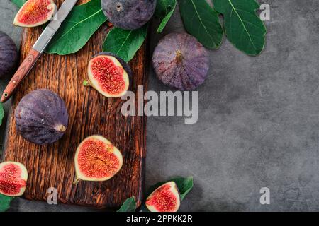 Figs cut into slices on a wooden cutting board next to an old knife. Figs surrounded by fig tree leaves, laid out on gray kitchen table. seasonal ripe Stock Photo