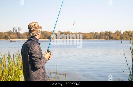 A male fisherman threw a spinning rod into a lake or river on a sunny day. Shore fishing. A fisherman on the river bank holds a spinning rod in his hand. The concept of country rest. Fishing article. Stock Photo