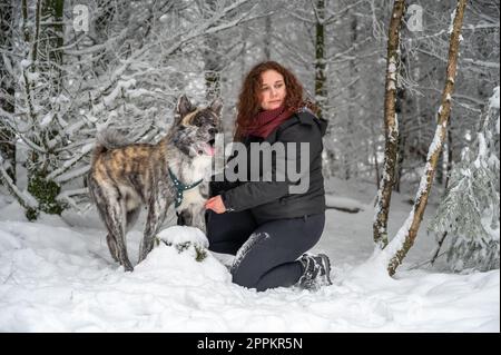 Young woman is kneeling next to her akita inu dog with gray fur, in the forest during winter with lots of snow Stock Photo