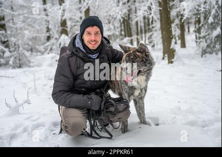 Master with his gray colored Akita Inu dog in the forest during winter with lots of snow, man is smiling and looking at camera while embracing his dog Stock Photo